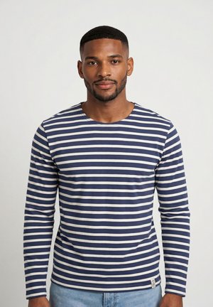 Uomo con barba curata che indossa una maglia a maniche lunghe a righe blu navy e bianche e jeans azzurri, in piedi davanti a uno sfondo chiaro e uniforme.