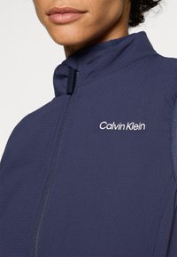 Casaco com fecho zip em azul-marinho e colarinho alto, apresentando uma textura subtil, logo em branco no lado esquerdo do peito e um design aerodinâmico.