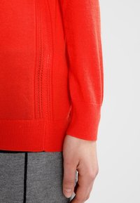 Gros plan sur la main d'une personne reposant à côté d'un pull rouge à manches longues avec des détails en maille, associé à un pantalon gris avec une couture verticale noire.