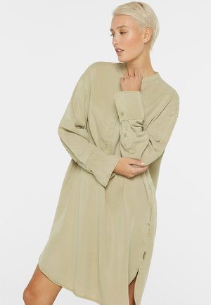 Frau mit kurzen blonden Haaren, die ein locker sitzendes, langärmliges beiges Hemdblusenkleid trägt und mit den Händen in der Nähe von Brust und Taille vor einem schlichten Hintergrund posiert.