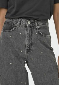 Jeans gris de cintura alta con pequeños tachuelas plateadas esparcidas en el frente, combinados con una blusa negra de manga corta metida por dentro.