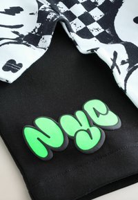 Tissu noir avec un logo "NYC" vert en relief. En dessous, un haut blanc avec un motif à carreaux noir et des graphismes abstraits.