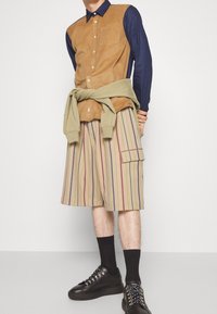 Brązowo-niebieska koszula z guzikiem, paskowe beżowe szorty w odcieniach czerwieni i niebieskiego, oliwkowy sweter zawiązany w talii, czarne sneakersy.