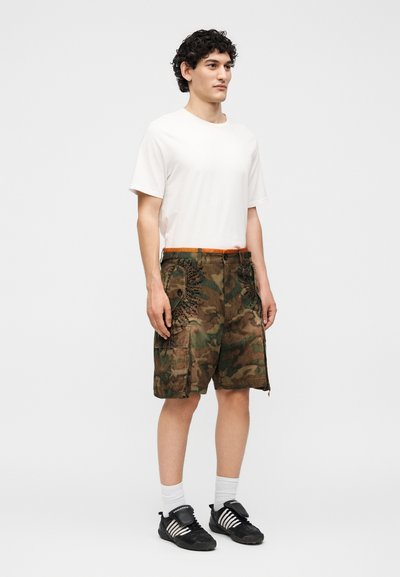 Άνδρας στέκεται φορώντας απλό λευκό t-shirt, καμουφλάζ cargo σορτς με μαύρες χάντρες, λευκές κάλτσες και μαύρα αθλητικά παπούτσια με λευκές ρίγες.