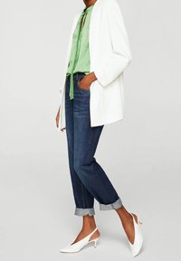 Blazer bianco, blusa verde a pois con cravatta, jeans in denim arrotolati e décolleté bianche con tacco basso. Design strutturato e sartoriale.
