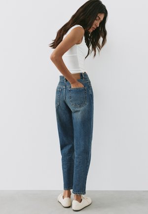 Vrouw met lang donker haar, een witte mouwloze top, blauwe high-waist jeans en witte sneakers, staand tegen een effen witte muur.