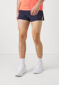 Mann trägt marineblaue Sportshorts, ein oranges Mesh-Oberteil, weiße Knöchelsocken und weiße Laufschuhe und steht auf einem weißen Boden.