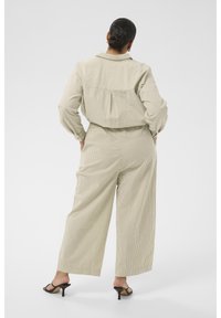 Femme aux cheveux noirs en chignon bas, portant un pantalon large beige à fines rayures et une chemise à manches longues assortie, debout, mains sur les hanches, dos face au spectateur.