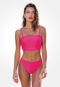 Frau mit lockigem Haar, die einen hellpinkfarbenen Zweiteiler-Badeanzug trägt, steht vor einem schlichten weißen Hintergrund und schaut geradeaus.