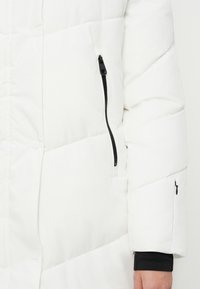 Hvid polstret jakkeærme med sort lynlås lomme og synlig del af håndleddet dækket af sort indre manchet.