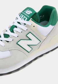 Světle šedé tenisky s bílými síťovinovými panely, zelenými akcenty, strukturovanou semišovou úpravou a výrazným bílým logem "N". Nabízí polstrovanou podporu kotníku a černou gumovou podrážku.