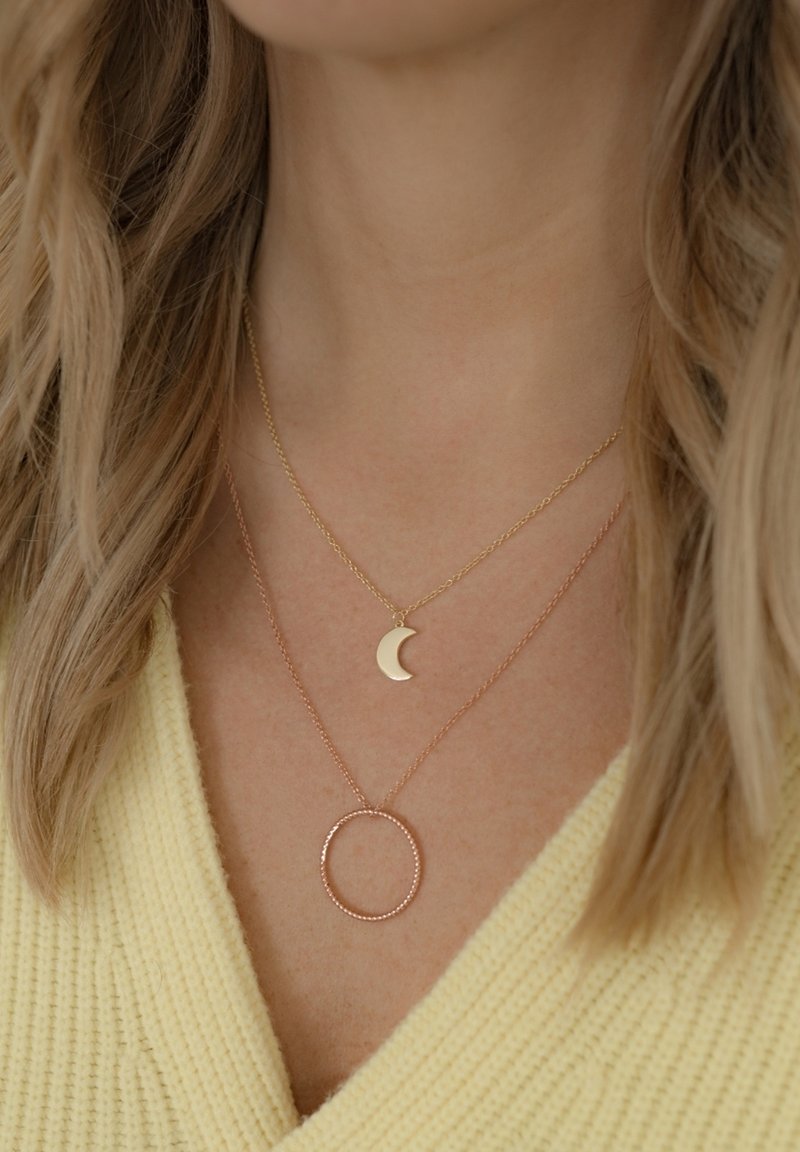 Slāņotas kaklarotas ar zelta pusmēness piekariņu un rozā zelta apļveida piekariņu uz smalkām ķēdēm. Tekstūrēts aizdare un ķēdes detaļas.