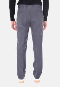 Uomo che indossa pantaloni grigi scuri e scarpe bianche, in piedi con le mani rilassate lungo i fianchi, visto da dietro su uno sfondo chiaro e semplice.