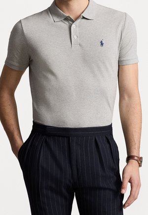 Mężczyzna w jasnoszarej koszulce polo z krótkim rękawem i trzema guzikami oraz granatowych spodniach w prążki, ręce w kieszeniach, na nadgarstku brązowy skórzany zegarek.