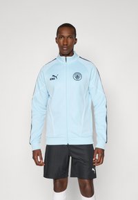 Light blue zip-up jacket with dark blue accents, Puma logo, and club emblem. Features ribbed cuffs, and trim, worn with dark shorts.