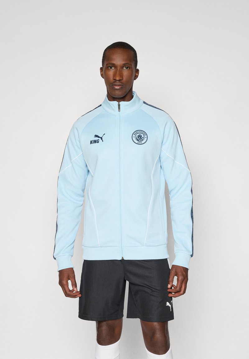 Light blue zip-up jacket with dark blue accents, Puma logo, and club emblem. Features ribbed cuffs, and trim, worn with dark shorts.