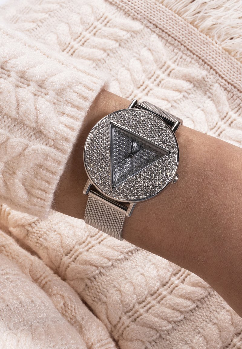 Ceas de mână cu brățară argintie din plasă și cadran rotund cu un triunghi acoperit cu cristale, purtat pe încheietura mâinii peste un pulover crem cu model împletit tip cablu.