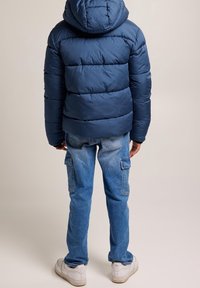 Mörkblå puffjacka med huva och sömnadsdetaljer, i kombination med ljusblå cargojackor och vita sneakers. Bakifrån.