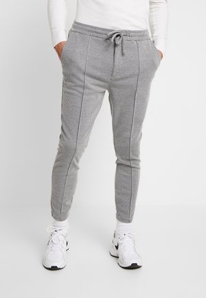Man draagt slim-fit joggingbroek met ruitpatroon en voorste naden, met trekkoord in de taille, witte sokken en witte Nike sneakers, handen in de zakken.