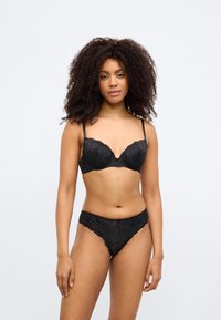 Čierna čipková podprsenka s vypchávkami a nastaviteľnými ramienkami, kombinovaná so zodpovedajúcimi čipkovanými nohavičkami s vlnitým okrajom a kvetinovým vzorom.