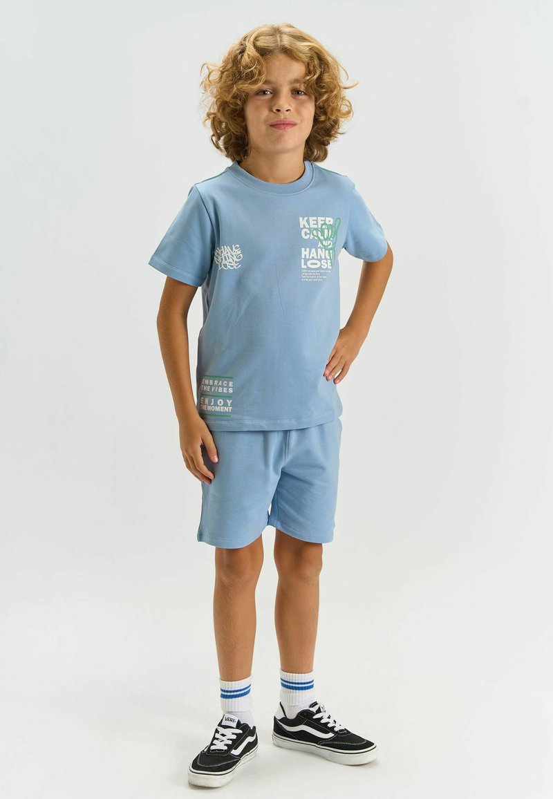 Garçon aux cheveux bouclés portant un t-shirt bleu clair et un short, debout avec une main sur la hanche, baskets noires avec des chaussettes blanches.