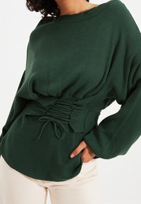 Grüner, gerippter Pullover mit lockerem Sitz, ausgestattet mit einem gerafften Bund und einem Kordelzug-Akzent. Lange Ärmel mit breiten Bündchen.