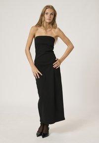 Melnas strapless maxi kleitas ar pieguļošu korpusu un taisnu siluetu. Gluda tekstūra un apakšmalu sasniedz potītes, kombinēta ar norādītām papēžiem.