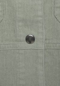 Lichtgroene corduroy stof met verticale ribbels, voorzien van een ronde metalen knop in het midden en twee opgestikte zakken.
