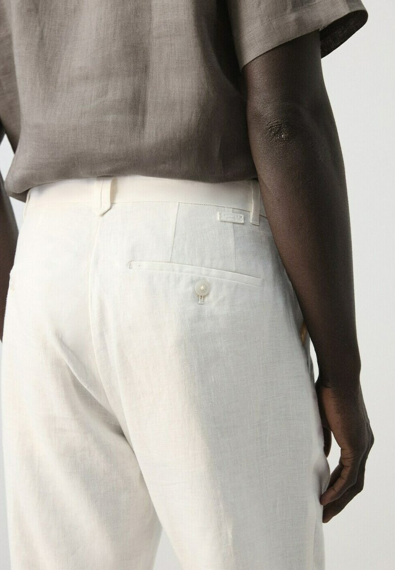 Personne portant un pantalon en lin beige clair avec une poche arrière boutonnée et une chemise à manches courtes taupe, vue de la taille vers le bas.