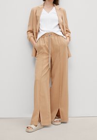 Pantaloni beige a gamba larga con spacchetti laterali, cintura elastica e cordoncino. Abbinati a una maglietta bianca a scollo a V e scarpe di colore chiaro.