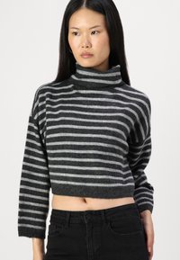 Maglione a collo alto a righe grigie e nere, design cropped, texture morbida, con colletto alto e spalle cadenti. Abbinato a jeans neri.