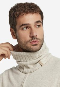 Lys beige, strikket turtleneck sweater med ribbet tekstur og en foldet krave. Har et lille vævet mærke nær halsudskæringen.
