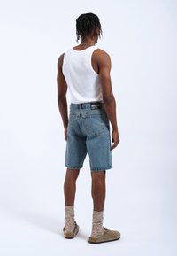 Shorts in denim blu con taglio dritto, orlo sfrangiato e tasche posteriori; abbinati a una canotta bianca e pantofole beige con calzini strutturati.
