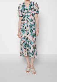 Mujer vistiendo un vestido midi floral en tonos pastel con mangas abullonadas y sandalias abiertas beige, de pie contra un fondo claro liso.