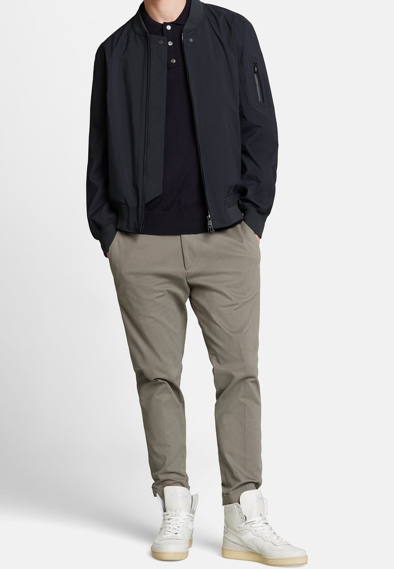 Blouson aviateur bleu marine avec col côtelé, chinos gris clair et baskets montantes blanches. La veste est dotée d'une fermeture éclair et d'une poche latérale.