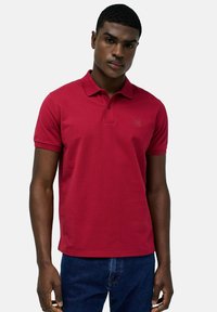 Polo rosso realizzato in cotone strutturato, con colletto standard, maniche corte e un piccolo logo del marchio sul petto. Abbinato a un jeans.
