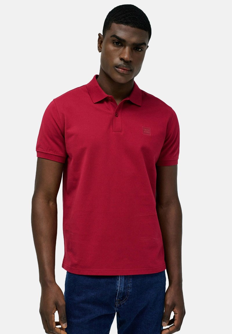 Polo rosso realizzato in cotone strutturato, con colletto standard, maniche corte e un piccolo logo del marchio sul petto. Abbinato a un jeans.