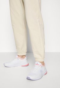 Zapatillas deportivas blancas con materiales de malla y sintéticos, con detalles en rosa y púrpura, cierre de cordones y cuello acolchado. Se ven pantalones beige.