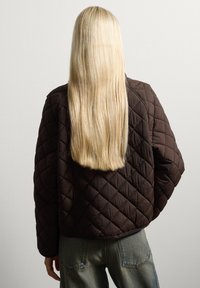 Braune gesteppte Jacke mit einem strukturierten Muster, runder Halslinie und überschnittenen Schultern, kombiniert mit hellblauen Jeans.