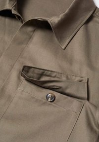 Chemise à manches courtes vert olive en tissu lisse, dotée d'une poche poitrine avec bouton et d'un col pointu, mettant en avant des lignes épurées et une simplicité élégante.