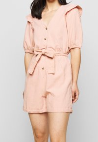 Ljusrosa kort jumpsuit med V-hals, puffärmar med volangdetaljer, midjebälte och knäppning fram.