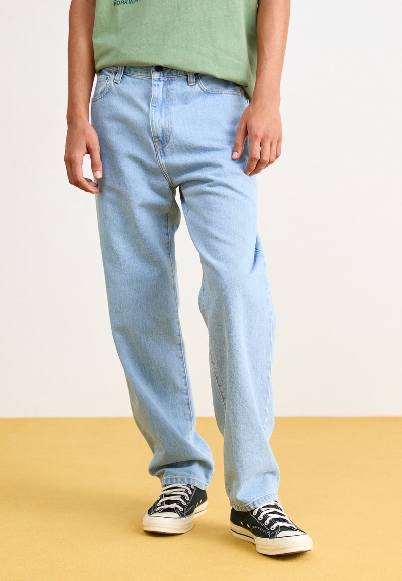 Jeans de mezclilla azul claro con un corte relajado, con cierre de botón y cremallera, pasadores para cinturón y costuras visibles. Llevado con zapatillas negras.