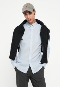 Light blue, pinstriped button-up shirt with a chest pocket, combined with a black sweater draped over the shoulders and a gray cap.