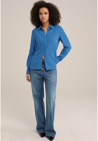 Camicia blu a maniche lunghe con colletto, abbinata a jeans svasati azzurri e décolleté nere a punta. Tessuto morbido.