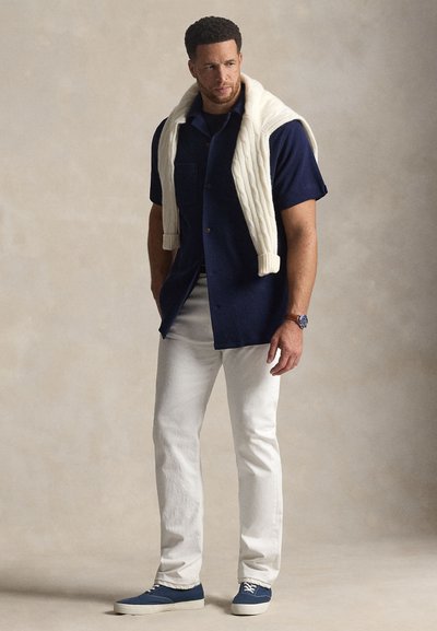 Μπλε πουκάμισο με κοντά μανίκια, λευκό πουλόβερ πλεκτό με καλώδια ριγμένο στους ώμους, λευκό παντελόνι, μπλε παπούτσια, στέκεται μπροστά σε ουδέτερο φόντο.