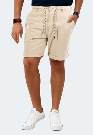 Beige cotton shorts with a slim fit, rolled hems, front pockets, and a patterned drawstring. Paired with white sneakers.