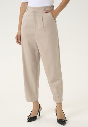 Beige skræddersyede bukser med høj talje, forreste plisseringer, sidelommer, kombineret med sorte slingback-hælspidse sko på en stående person.