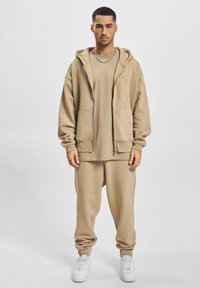Sweat-shirt à capuche beige avec poches avant, associé à un jogging assorti. Tissu doux, coupe décontractée et poignets élastiques. Des baskets blanches complètent la tenue.