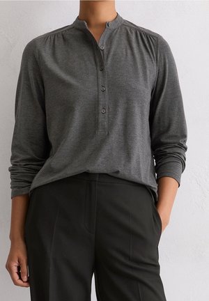 Person, die ein dunkelgraues Langarm-Henley-Shirt mit Knöpfen trägt, in hoch taillierte schwarze Hose gesteckt, steht vor einer hellen Wand.