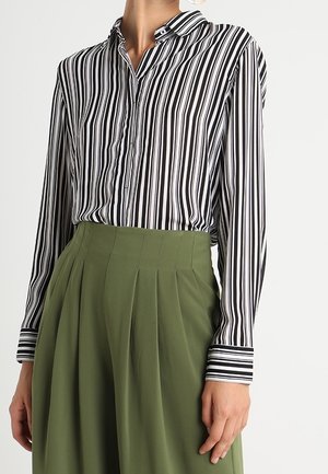 Mujer con una camisa de manga larga con rayas verticales en blanco y negro metida en una falda plisada de talle alto de color verde oliva.